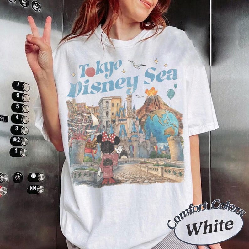Pu&ograve; includere: T-shirt bianca con grafica "Tokyo Disney Sea" in testo blu e rosso. Il design include illustrazioni di un castello, Minnie Mouse e un globo. La maglietta &egrave; etichettata "Comfort Colors White".