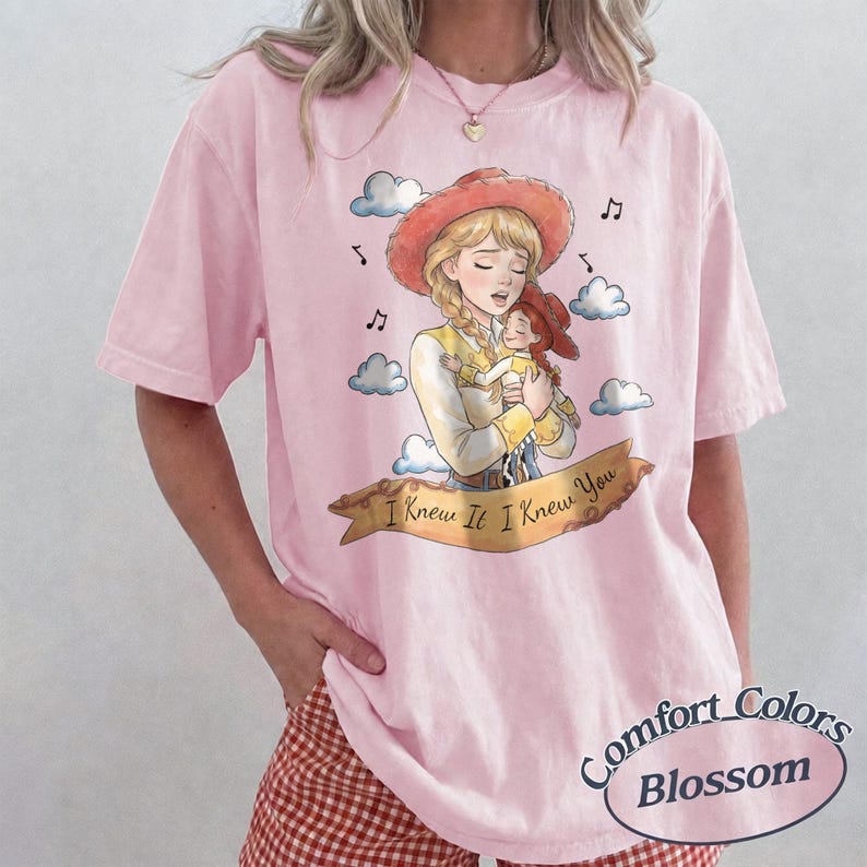 May include: A light pink t-shirt featuring a graphic illustration of a blonde character in a cowgirl hat holding a smaller doll version of herself. The design includes blue clouds, musical notes, and a banner with the text I Knew It I Knew You.