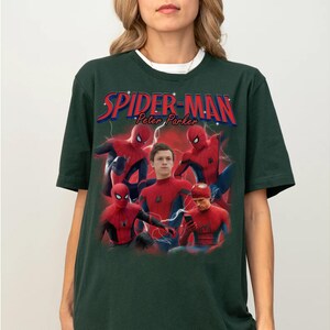 Może przedstawiać: Ciemnozielona koszulka z napisem "Spider-Man Peter Parker" w kolorze czerwonym i niebieskim. Koszulka przedstawia wiele zdjęć Spider-Mana w pozach akcji. Ma biały okrągły kołnierz.