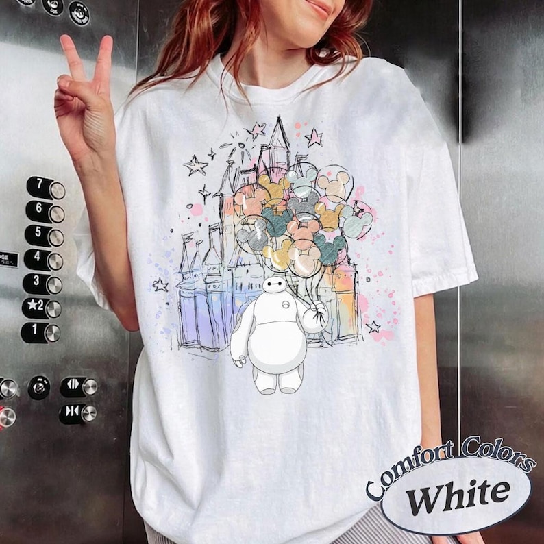 Puede incluir: Camiseta blanca Comfort Colors con una colorida ilustraci&oacute;n de Baymax sosteniendo globos de Mickey Mouse frente a un castillo. El dise&ntilde;o incluye estrellas y salpicaduras pastel. El texto "Comfort Colors White" est&aacute; impreso abajo a la derecha.
