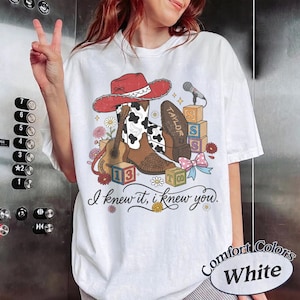 Pu&ograve; includere: Una t-shirt bianca con una stampa grafica di un cappello da cowboy rosso, uno stivale da cowboy marrone e bianco con stampa muccata, una chitarra acustica, un microfono, cubi dell'alfabeto e un fiocco. Il testo recita "TAYLOR", "13" e "I knew it, i knew you."