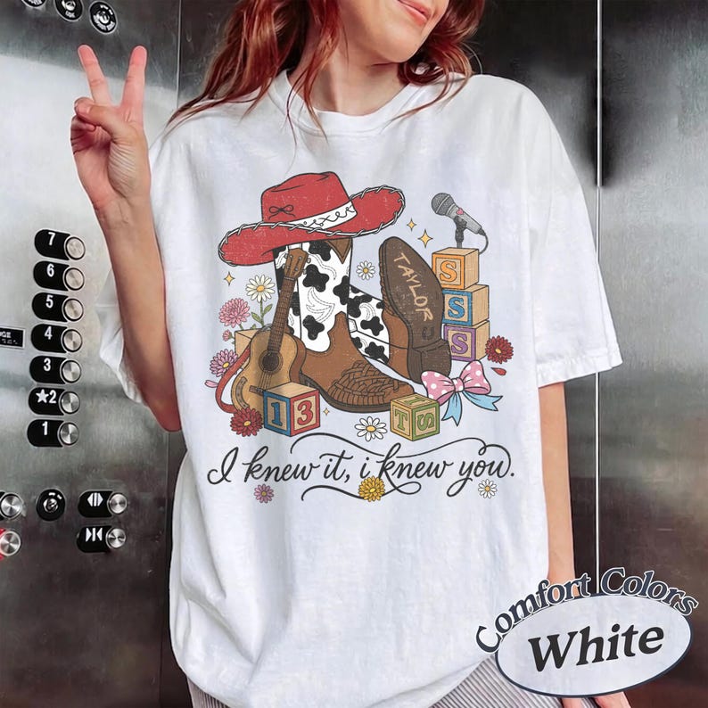 Pu&ograve; includere: Una t-shirt bianca con una stampa grafica di un cappello da cowboy rosso, uno stivale da cowboy marrone e bianco con stampa muccata, una chitarra acustica, un microfono, cubi dell'alfabeto e un fiocco. Il testo recita "TAYLOR", "13" e "I knew it, i knew you."