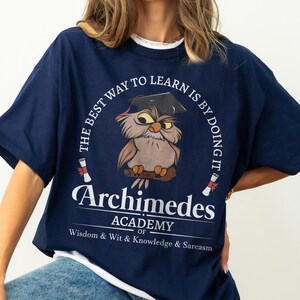 May include: A navy blue t-shirt featuring a graphic of an owl wearing a graduation cap, perched on a branch. The text reads: THE BEST WAY TO LEARN IS BY DOING IT, Archimedes ACADEMY OF Wisdom & Wit & Knowledge & Sarcasm, with two diploma scrolls.
