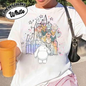Puede incluir: Camiseta blanca con una ilustraci&oacute;n de dibujos animados de Baymax sosteniendo globos de Mickey Mouse frente a un castillo. La camiseta tiene las palabras "Comfort Colors" y "White" impresas. El dise&ntilde;o incluye estrellas y acentos coloridos.
