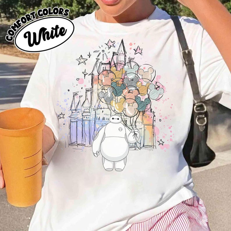 Puede incluir: Camiseta blanca con una ilustraci&oacute;n de dibujos animados de Baymax sosteniendo globos de Mickey Mouse frente a un castillo. La camiseta tiene las palabras "Comfort Colors" y "White" impresas. El dise&ntilde;o incluye estrellas y acentos coloridos.