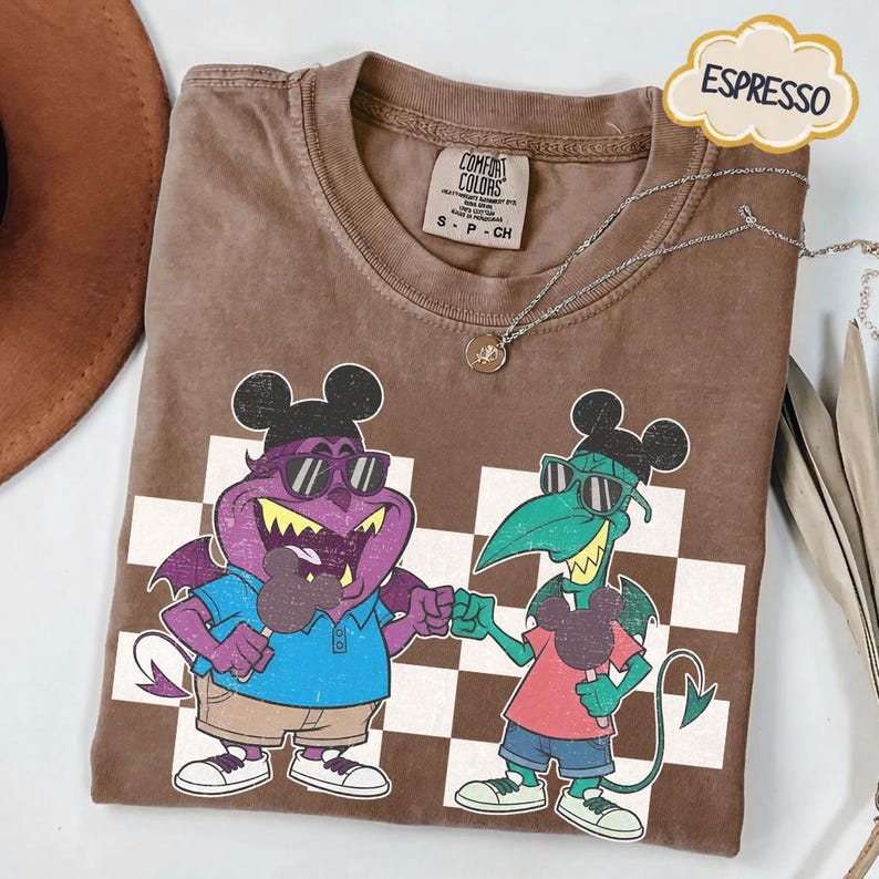 May include: Espresso-colored t-shirt with a cartoon graphic of two monster characters wearing sunglasses and Mickey Mouse ears, fist-bumping. The shirt has a checkered pattern and a Comfort Colors tag. A silver necklace with a pendant is draped across the shirt.