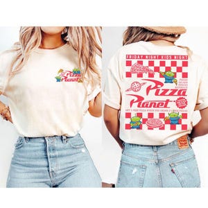 以下が含まれることがあります： 前面にPizza Planetのロゴとエイリアンキャラクターがプリントされたクリーム色のTシャツ。背中には「Friday Night Kids Night」と「Pizza Planet」の文字が赤色でプリントされた赤と白のチェック柄のデザイン。