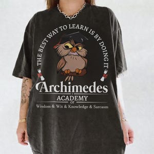 May include: A charcoal gray t-shirt featuring a graphic of an owl wearing a graduation cap. The text reads: THE BEST WAY TO LEARN IS BY DOING IT, Archimedes ACADEMY OF WISDOM & WIT & KNOWLEDGE & SARCASM. Two small diploma scrolls are also printed.