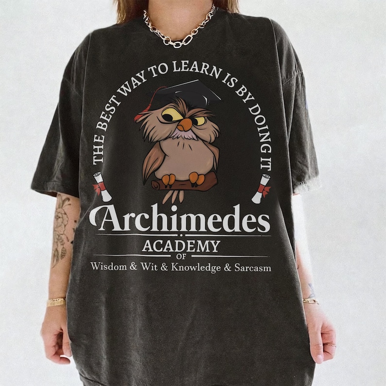 May include: A charcoal gray t-shirt featuring a graphic of an owl wearing a graduation cap. The text reads: THE BEST WAY TO LEARN IS BY DOING IT, Archimedes ACADEMY OF WISDOM & WIT & KNOWLEDGE & SARCASM. Two small diploma scrolls are also printed.