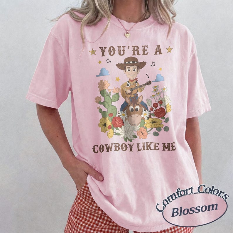 Op de afbeelding: Een lichtroze T-shirt met korte mouwen en een grafische print van een cartoon-cowboy die gitaar speelt op een paard, omringd door cactussen en bloemen. De tekst "YOU'RE A" en "COWBOY LIKE ME" is in het bruin boven en onder de afbeelding gedrukt.