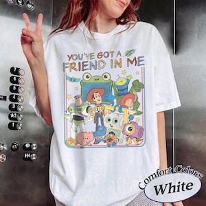 May include: A white short-sleeve t-shirt featuring a colorful graphic print of various animated characters from the Toy Story franchise. The text "YOU'VE GOT A FRIEND IN ME" is printed in a multi-colored font above the character illustration.