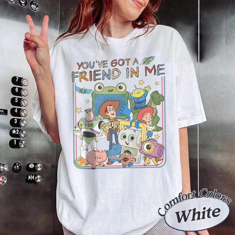 May include: A white short-sleeve t-shirt featuring a colorful graphic print of various animated characters from the Toy Story franchise. The text "YOU'VE GOT A FRIEND IN ME" is printed in a multi-colored font above the character illustration.