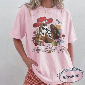 Pu&ograve; includere: Una t-shirt rosa chiaro con una stampa grafica raffigurante un cappello da cowboy rosso, uno stivale marrone e bianco con stampa mucca, una chitarra acustica, blocchi dell'alfabeto e un microfono. Il testo recita: I knew it, i knew you. Include fiori.