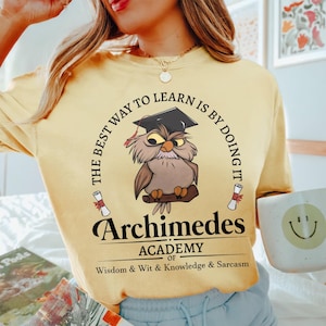 May include: A pale yellow t-shirt featuring a graphic of a brown owl wearing a black graduation cap. The text reads: THE BEST WAY TO LEARN IS BY DOING IT, Archimedes ACADEMY OF WISDOM & WIT & KNOWLEDGE & SARCASM, with two small scrolls.