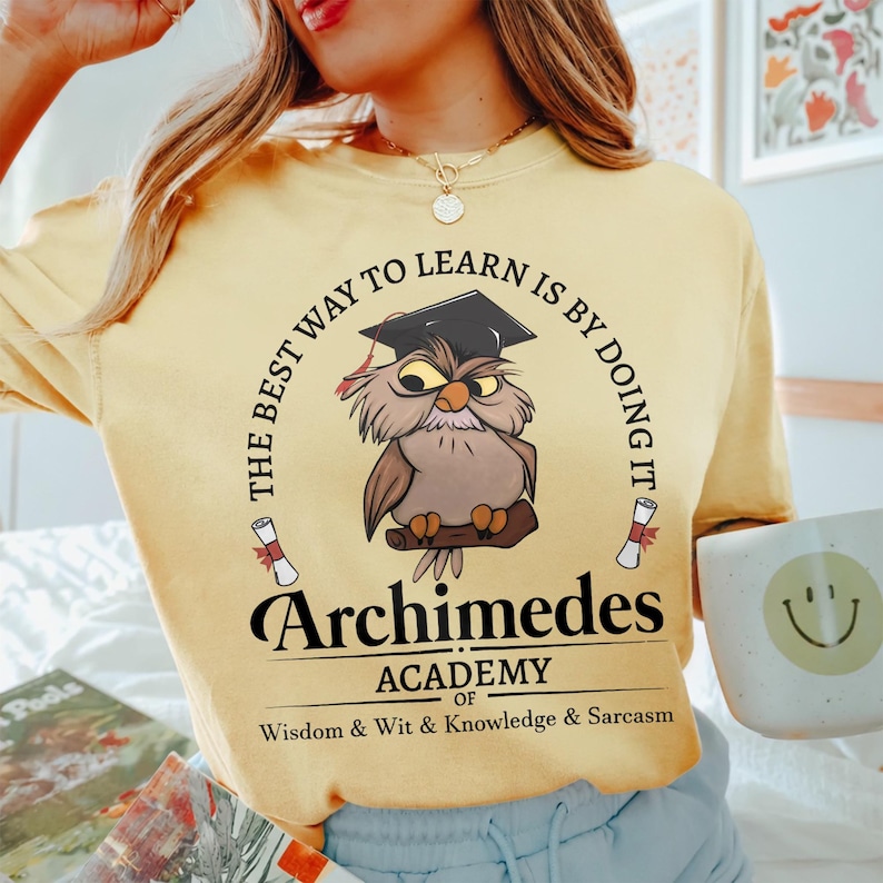 May include: A pale yellow t-shirt featuring a graphic of a brown owl wearing a black graduation cap. The text reads: THE BEST WAY TO LEARN IS BY DOING IT, Archimedes ACADEMY OF WISDOM & WIT & KNOWLEDGE & SARCASM, with two small scrolls.