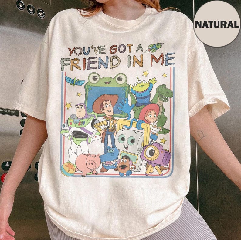May include: A natural-colored t-shirt featuring a graphic print of various animated toy characters. The text "YOU'VE GOT A FRIEND IN ME" is printed above the characters. The design includes a cowboy, space ranger, dinosaur, and other figures.