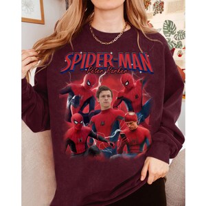 Może przedstawiać: Bordowa bluza z grafiką Spider-Mana i napisem "Spider-Man Peter Parker" w kolorze czerwonym i niebieskim. Projekt zawiera wiele zdjęć Spider-Mana w pozach akcji, ze zdjęciem aktora w centrum.