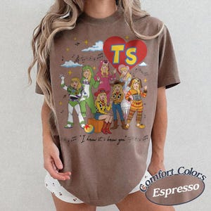 May include: A brown short-sleeve t-shirt featuring a graphic of seven cartoon characters dressed in various costumes. Above the characters is a red heart with the yellow text TS. Below the characters is the text I knew it, I knew you.