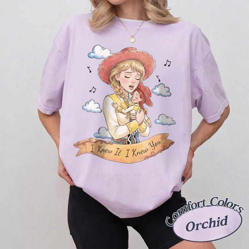 May include: A light purple short-sleeve t-shirt featuring a graphic illustration of a blonde cowgirl holding a smaller cowgirl doll. The design includes clouds, musical notes, and a banner with the text I Knew It I Knew You.