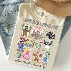 May include: Cream-colored t-shirt featuring a colorful "Universal Studios" graphic with various cartoon characters and the text "Where Adventure Comes to Life. 2026". The shirt is folded on top of blue jeans.