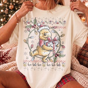 May include: A cream-colored t-shirt featuring a graphic of Winnie the Pooh wearing a Santa hat and holding a gift. The design includes a circular string of Christmas lights, holly leaves, snowflakes, and the repeated text ho ho ho.