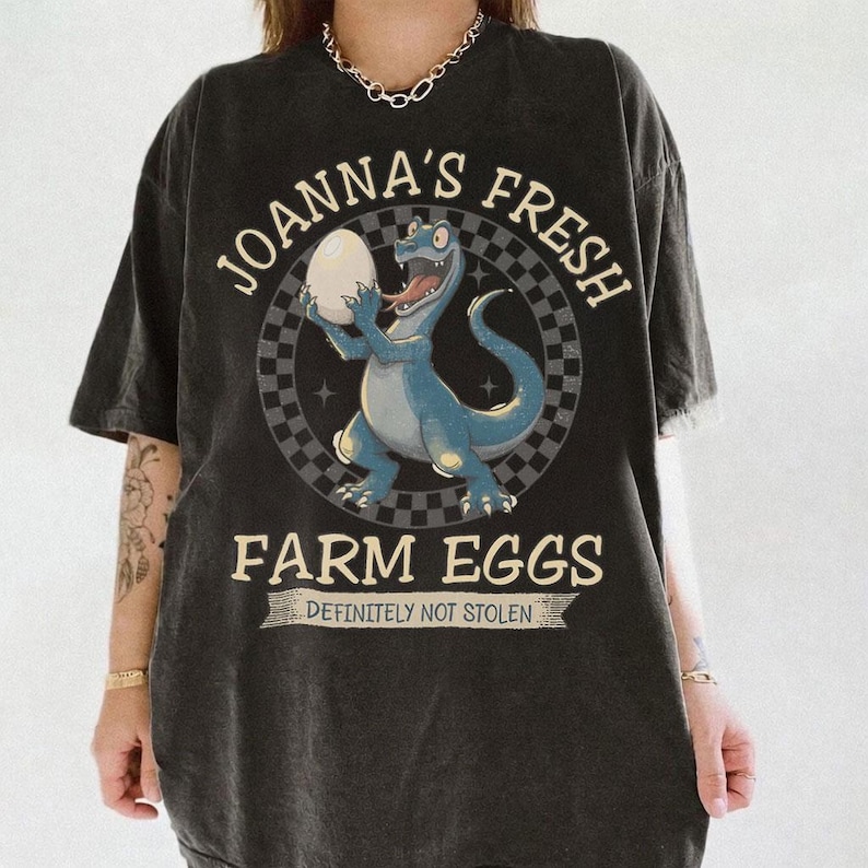 May include: Dark gray t-shirt featuring a cartoon dinosaur holding an egg, with the text "JOANNA'S FRESH FARM EGGS" and "DEFINITELY NOT STOLEN". The dinosaur is blue with a red tongue, set against a checkered background.