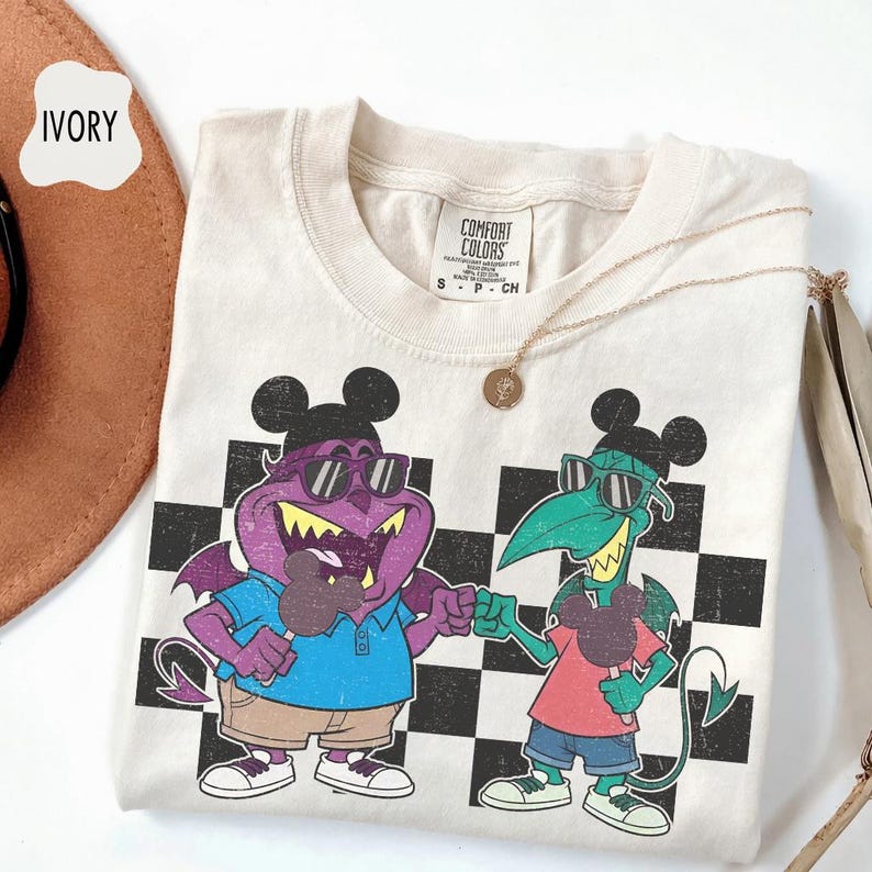 May include: Ivory-colored t-shirt featuring a cartoon graphic of two devilish characters wearing sunglasses and Mickey Mouse ears, set against a black and white checkered background. The shirt has a crew neck and short sleeves. A gold necklace is draped across the shirt.