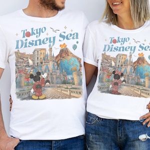 Pu&ograve; includere: T-shirt bianche con una grafica colorata raffigurante Topolino e Minnie Mouse in abiti tradizionali giapponesi, con la scritta "Tokyo Disney Sea". Il design include un castello, un globo e altri elementi del parco.