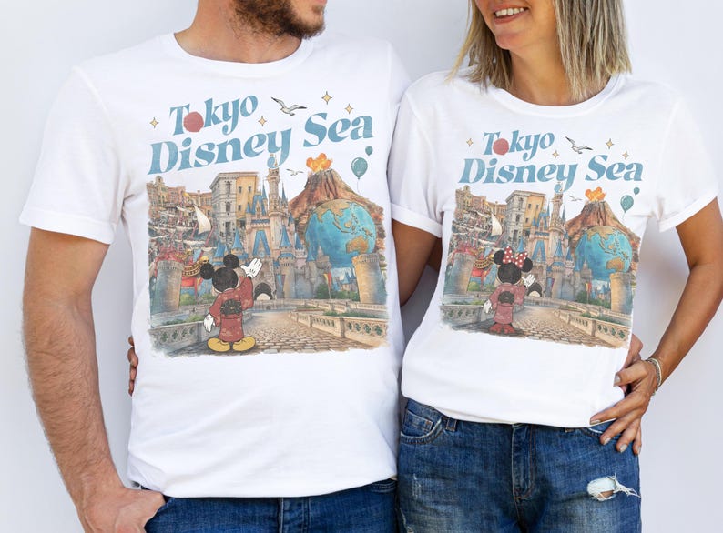 Pu&ograve; includere: T-shirt bianche con una grafica colorata raffigurante Topolino e Minnie Mouse in abiti tradizionali giapponesi, con la scritta "Tokyo Disney Sea". Il design include un castello, un globo e altri elementi del parco.