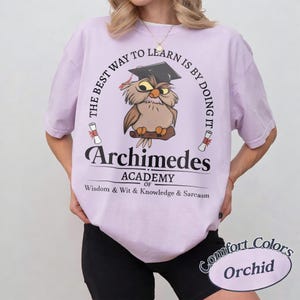 May include: A light purple t-shirt featuring a graphic of an owl wearing a graduation cap. The text reads: THE BEST WAY TO LEARN IS BY DOING IT, Archimedes ACADEMY OF WISDOM & WIT & KNOWLEDGE & SARCASM. Two small diploma scrolls flank the owl.