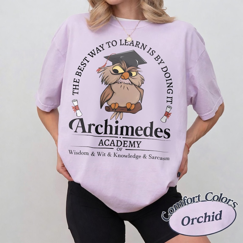 May include: A light purple t-shirt featuring a graphic of an owl wearing a graduation cap. The text reads: THE BEST WAY TO LEARN IS BY DOING IT, Archimedes ACADEMY OF WISDOM & WIT & KNOWLEDGE & SARCASM. Two small diploma scrolls flank the owl.