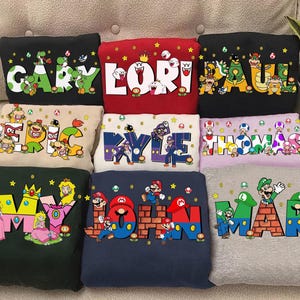 May include: A collection of nine sweatshirts in various colours, each featuring a large name printed in block letters. The letters are decorated with different video game characters, including Mario, Luigi, Princess Peach, Bowser, and various enemies.