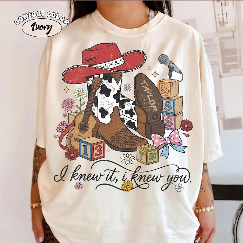 Pu&ograve; includere: Una t-shirt color avorio con una stampa grafica di un cappello da cowboy rosso, uno stivale con stampa mucca marrone e bianca, una chitarra acustica, un microfono, blocchi dell'alfabeto e fiori. Il testo recita "I knew it, i knew you" e "TAYLOR".