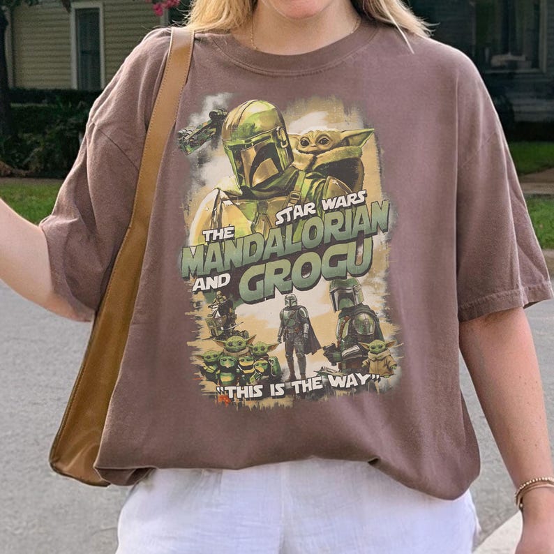 Może przedstawiać: Brązowa koszulka z grafiką Star Wars z The Mandalorian i Grogu. Projekt zawiera obrazy Mandalorianina, Grogu i innych postaci z napisem "THIS IS THE WAY".