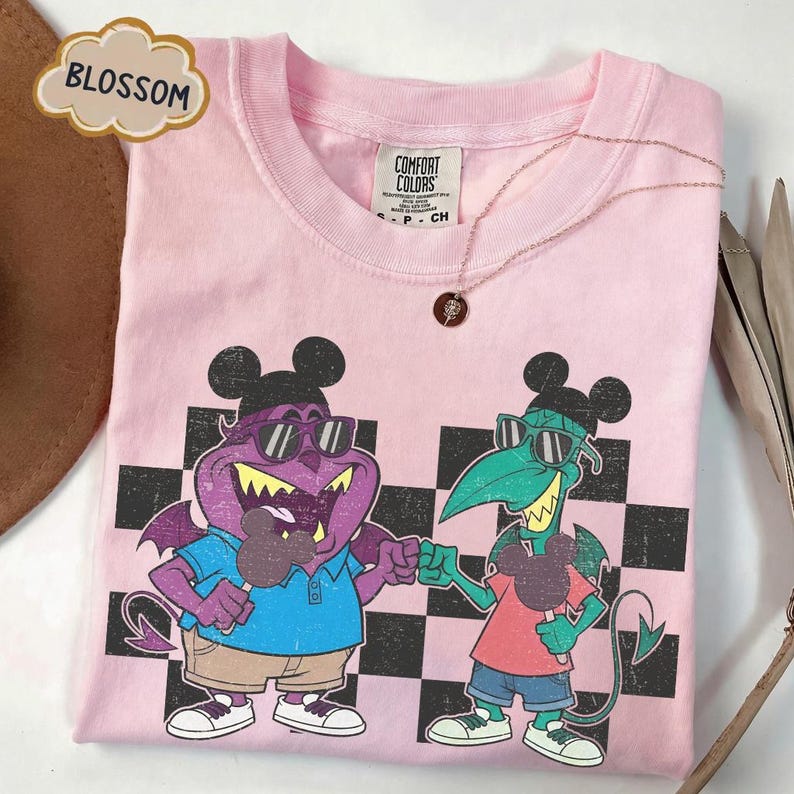 May include: Pink Comfort Colors t-shirt featuring a cartoon graphic of two monster characters wearing sunglasses and holding ice cream. The shirt has a black and white checkered pattern and a rose gold necklace. The word "BLOSSOM" is in a cloud-shaped text box.