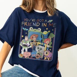 May include: A navy blue t-shirt featuring a graphic print of various animated characters from the Toy Story franchise. The text "YOU'VE GOT A FRIEND IN ME" is printed in a colorful, textured font above the group of characters, which includes Woody, Buzz Lightyear, Jessie, Rex, and others.
