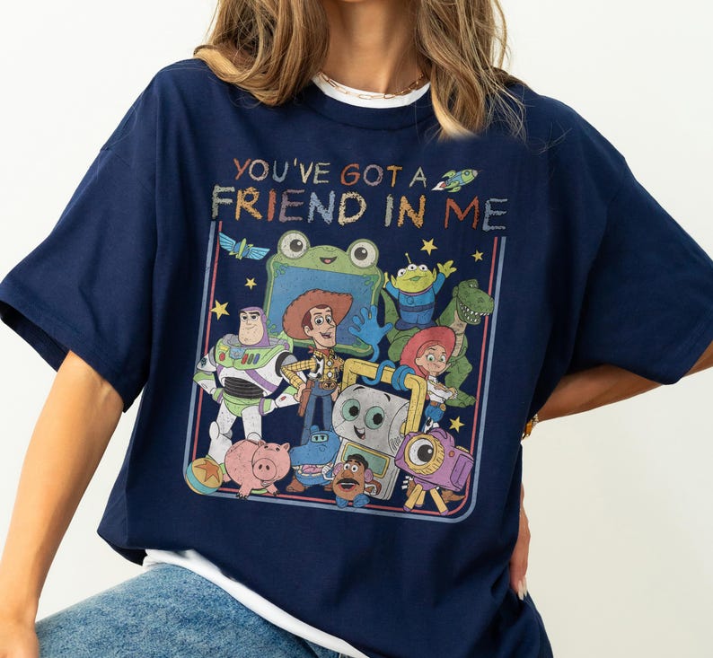 May include: A navy blue t-shirt featuring a graphic print of various animated characters from the Toy Story franchise. The text "YOU'VE GOT A FRIEND IN ME" is printed in a colorful, textured font above the group of characters, which includes Woody, Buzz Lightyear, Jessie, Rex, and others.