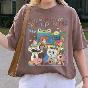May include: A brown short-sleeve t-shirt featuring a graphic print of Toy Story characters including Woody, Buzz Lightyear, Jessie, Rex, and Mr. Potato Head. The text "YOU'VE GOT A FRIEND IN ME" is printed above the colorful cartoon character illustration.