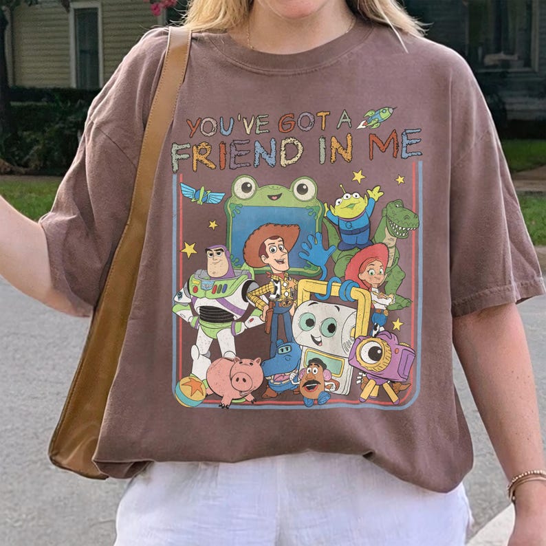 May include: A brown short-sleeve t-shirt featuring a graphic print of Toy Story characters including Woody, Buzz Lightyear, Jessie, Rex, and Mr. Potato Head. The text "YOU'VE GOT A FRIEND IN ME" is printed above the colorful cartoon character illustration.