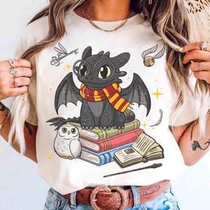 Puede incluir: Camiseta color crema con un dragón de dibujos animados con una bufanda, sentado sobre libros con un búho. El diseño incluye una llave, una snitch dorada y una varita. El dragón tiene ojos grandes y alas. Los libros son de varios colores.