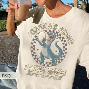 May include: Ivory-colored t-shirt featuring a cartoon dinosaur holding an egg, with the text "JOANNA'S FRESH FARM EGGS DEFINITELY NOT STOLEN" in a circular design. The dinosaur is blue with a red tongue.