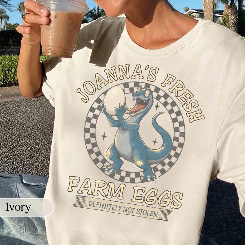 May include: Ivory-colored t-shirt featuring a cartoon dinosaur holding an egg, with the text "JOANNA'S FRESH FARM EGGS DEFINITELY NOT STOLEN" in a circular design. The dinosaur is blue with a red tongue.