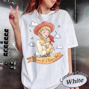 May include: A white t-shirt featuring a watercolour-style illustration of a blonde character in a red cowgirl hat holding a smaller doll. A tan banner across the bottom displays the text I Knew It I Knew You, surrounded by clouds and musical notes.
