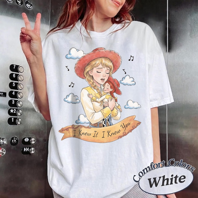 May include: A white t-shirt featuring a watercolour-style illustration of a blonde character in a red cowgirl hat holding a smaller doll. A tan banner across the bottom displays the text I Knew It I Knew You, surrounded by clouds and musical notes.