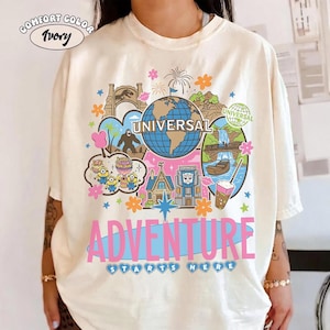 Könnte beinhalten: Elfenbeinfarbenes Comfort Color T-Shirt mit einem farbenfrohen Grafikdesign. Das Design enthält den Text "UNIVERSAL" auf einem Globus, Minions, einen Sasquatch und den Text "ADVENTURE STARTS HERE".