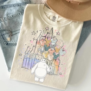 Puede incluir: Camiseta color crema con una ilustraci&oacute;n caprichosa de Baymax sosteniendo globos de Mickey Mouse frente a un castillo. El dise&ntilde;o incluye estrellas y acentos coloridos. La camiseta es de Comfort Colors.