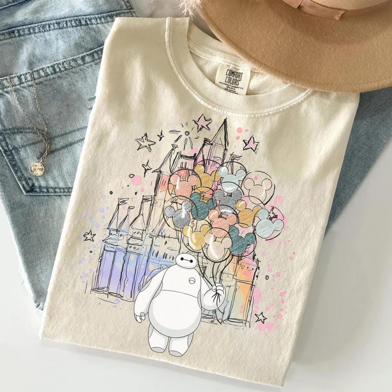 Puede incluir: Camiseta color crema con una ilustraci&oacute;n caprichosa de Baymax sosteniendo globos de Mickey Mouse frente a un castillo. El dise&ntilde;o incluye estrellas y acentos coloridos. La camiseta es de Comfort Colors.