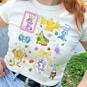 Puede incluir: Camiseta color crema con insectos de dibujos animados coloridos y el texto "bug's life" en amarillo. El diseño incluye varios personajes de insectos, flores y marcos decorativos. La camiseta es de estilo ajustado.