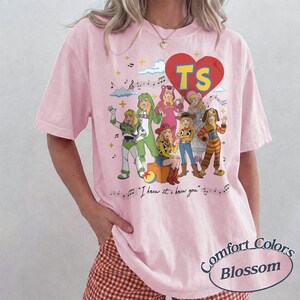 May include: A light pink short-sleeve t-shirt featuring a graphic print of various characters dressed in costumes. Above the characters is a red heart with the letters TS. Below the characters is the text I knew it, I knew you with musical notes.