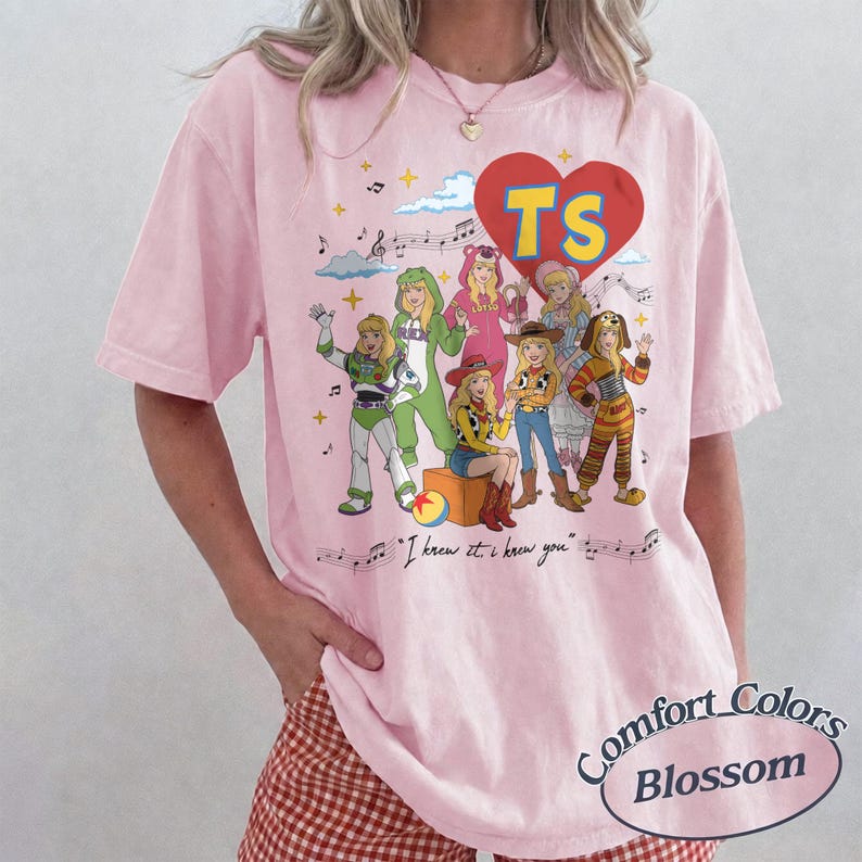 May include: A light pink short-sleeve t-shirt featuring a graphic print of various characters dressed in costumes. Above the characters is a red heart with the letters TS. Below the characters is the text I knew it, I knew you with musical notes.
